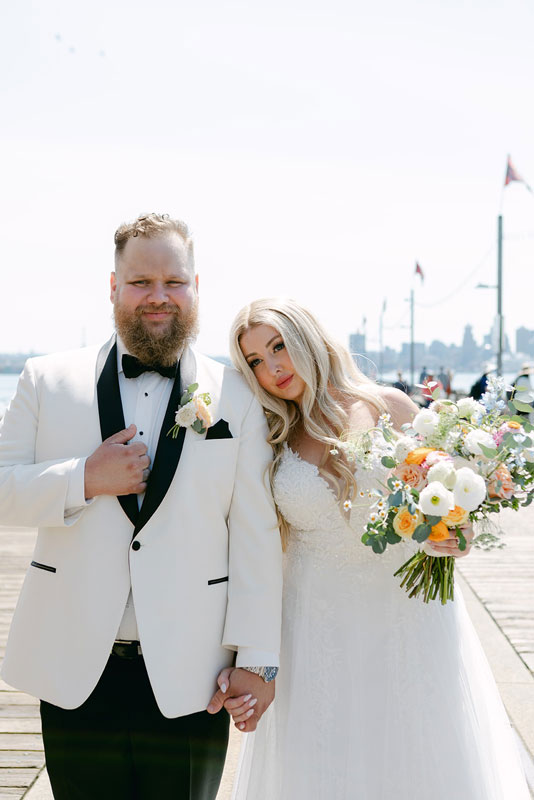 Bride with long blonde Hairalicious wig holding a bouquet beside her groom in a white tuxedo on a warfront dock