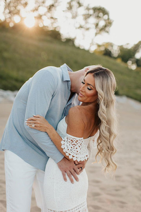 Bride with long wavy blonde Hairalicious wig embraced by her partner on a beach at sunset
